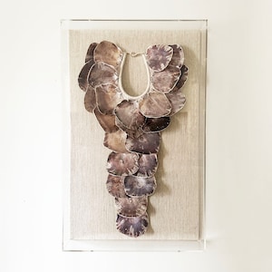 May include: A framed necklace made of brown, natural shell pieces. The necklace is displayed on a beige linen background.