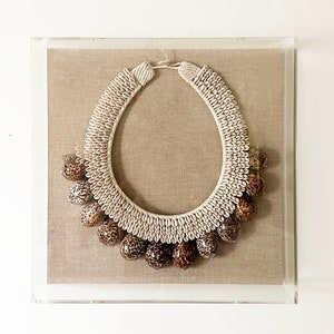 May include: A white and brown shell necklace with a large, round pendant. The necklace is displayed in a clear acrylic frame on a beige linen background.