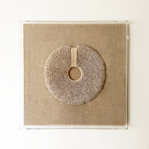 May include: A circular wall hanging made of woven natural materials, framed in a clear acrylic frame. The hanging has a central hole and is a light beige color.