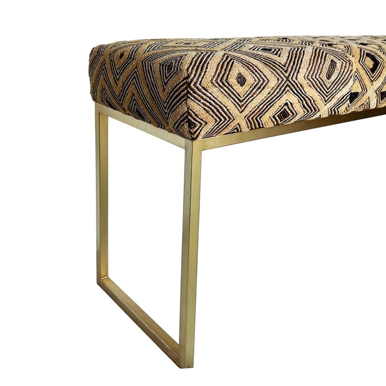 May include: A modern bench with a rectangular seat featuring a geometric pattern in shades of brown and gold. The bench has a gold-colored metal frame with square legs, creating a stylish and contemporary look.