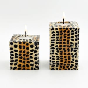 May include: Two square candle holders with a black, brown, and white animal print design. The candle holders are made of a textured material and have a small opening in the center for a tea light candle.