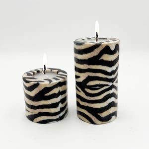 May include: Two lit pillar candles with a zebra stripe pattern in black and beige. The candles are different heights, with one short and wide, and the other tall and cylindrical. The flames are white.