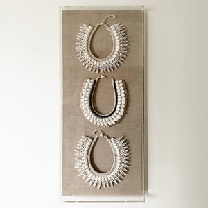 May include: Three white shell necklaces displayed in a clear acrylic frame on a beige linen background. The necklaces are made of different types of shells and are arranged in a vertical row.