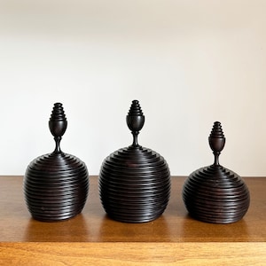May include: Three wooden decorative boxes with a dark brown finish. Each box has a rounded top with a layered, ribbed design and a small, pointed finial.
