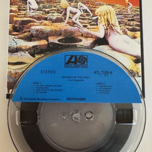 May include: A vintage reel-to-reel tape of Led Zeppelin's "Houses of the Holy" album. The album cover features a surreal image of nude figures on a rock formation under an orange sky. The tape reel is blue with white text.