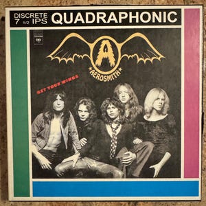 May include: Album cover for Aerosmith's "Get Your Wings." The cover features a black background with the band's name and logo in gold. The band members are pictured in a group photo. The top of the cover reads "DISCRETE QUADRAPHONIC."