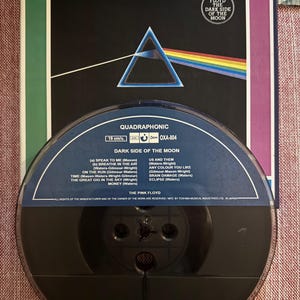 May include: A quadraphonic audio tape reel and album cover for Pink Floyd's "Dark Side of the Moon." The cover features a prism refracting light into a rainbow. The reel is black with track listings.