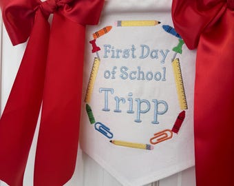 May include: White fabric banner with red, yellow, blue and green embroidered school supplies, including pencils, crayons and paper clips. The banner reads "First Day of School Tripp". The banner is hanging from a white string and is decorated with two red satin bows.