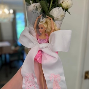 May include: A bouquet of white peonies wrapped in a decorative paper and white ribbon. A ballerina doll in a pink dress and tiara is attached to the bouquet. The white ribbon has pink embroidered ballet shoes and initials.