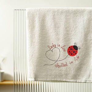 May include: Cream-coloured bath towel with embroidered ladybug and text "Lucky in Love Spotted in Life". The ladybug is red with black spots and a black head. The text is in a cursive font.