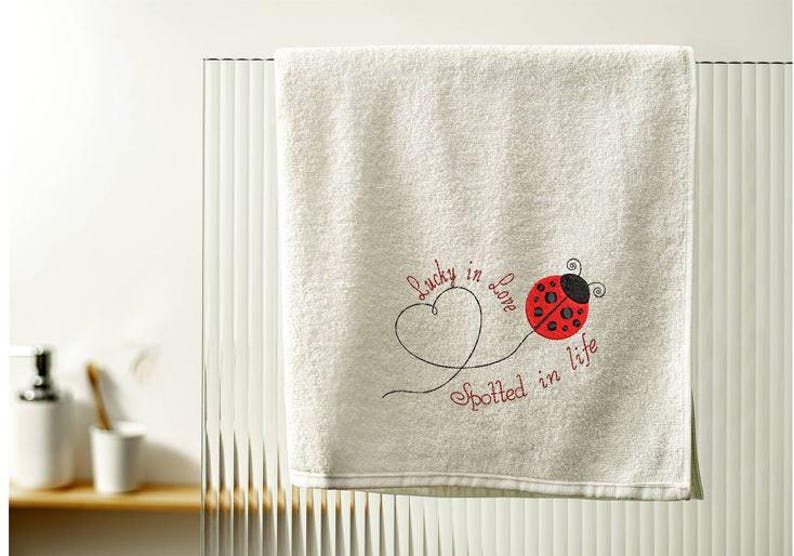 May include: Cream-coloured bath towel with embroidered ladybug and text "Lucky in Love Spotted in Life". The ladybug is red with black spots and a black head. The text is in a cursive font.