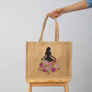 May include: A tan jute tote bag with a black silhouette of a person wearing a dress made of pink and purple roses. The bag has two tan handles and is held by a person.