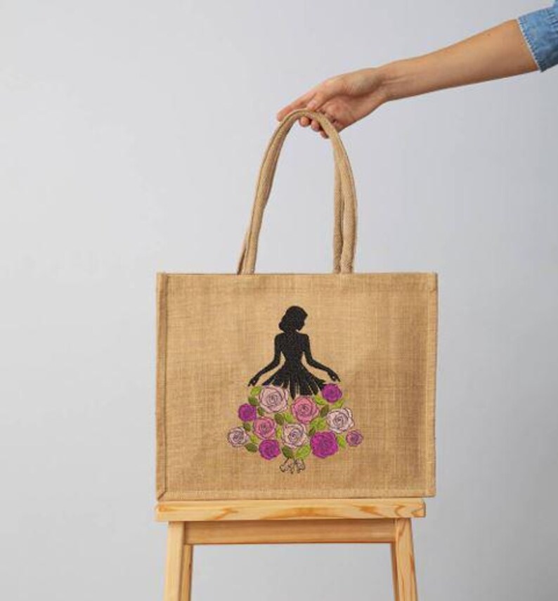 May include: A tan jute tote bag with a black silhouette of a person wearing a dress made of pink and purple roses. The bag has two tan handles and is held by a person.