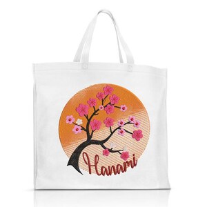 May include: White tote bag with an embroidered design of a pink cherry blossom tree in front of an orange sun. The text "Hanami" is embroidered below the tree.