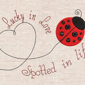 May include: Embroidered design of a red ladybird with black spots and a black head. A black heart-shaped line connects to the ladybird. Text reads "Lucky in Love Spotted in Life" in cursive on a beige background.