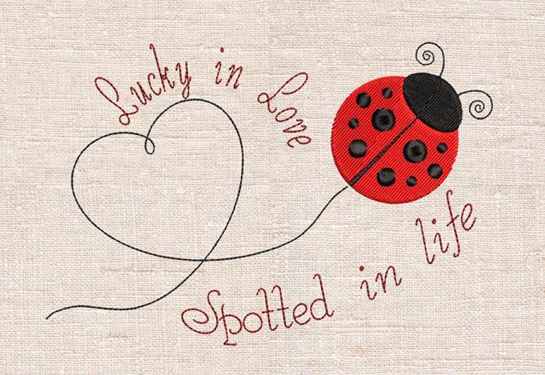 May include: Embroidered design of a red ladybird with black spots and a black head. A black heart-shaped line connects to the ladybird. Text reads "Lucky in Love Spotted in Life" in cursive on a beige background.