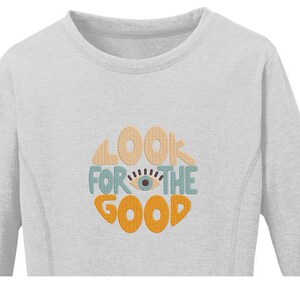 Pu&ograve; includere: Una felpa grigia chiaro a maniche lunghe con il testo "Look for the good" in un cerchio con un occhio stilizzato al centro. Il testo &egrave; in un font retr&ograve; con colori arancio, giallo e blu.