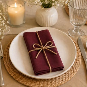 May include: A deep burgundy cloth napkin folded into a rectangle and tied with a natural jute twine bow, centred on a round off-white ceramic plate. The plate rests on a circular woven rattan placemat, accompanied by a silver fork, knife, and spoon.