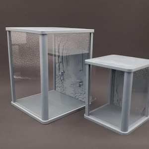 Puede incluir: Dos vitrinas cuadradas con marcos de color gris claro y paneles de vidrio transparente texturizado. La vitrina más grande presenta un diseño en relieve de un sendero forestal con setas en el panel posterior, junto a una vitrina más pequeña.