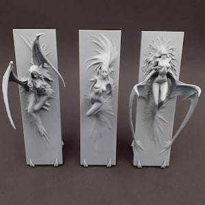 Peut inclure: Trois sculptures grises de figures féminines ailées, chacune montée sur une base rectangulaire. Les figures ont des ailes détaillées et des cheveux fluides, avec un thème fantastique ou gothique. Les sculptures sont exposées sur un fond neutre.