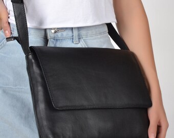 Op de afbeelding: Een zwarte leren schoudertas met een klep sluiting. De tas wordt gedragen over de schouder van een persoon die een wit shirt en een lichtblauwe denim short draagt.