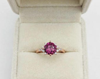 Puede incluir: Un anillo de oro rosa con una piedra central redonda de color rosa intenso sujeta por seis garras, flanqueada por tres pequeñas piedras transparentes a cada lado, presentado dentro de un joyero de color beige claro forrado de terciopelo.