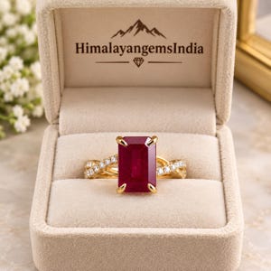 May include: A gold ring featuring a large, rectangular, deep red ruby center stone held by four prongs. The band has a twisted design encrusted with small, clear, round-cut diamonds. The ring is displayed in a beige box labeled HimalayangemsIndia.