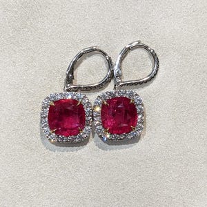 May include: A pair of drop earrings featuring cushion-cut red gemstones set in silver-toned metal. Each central stone is surrounded by a halo of small, clear, sparkling stones. The earrings have hinged lever-back closures with pavé-set accents.