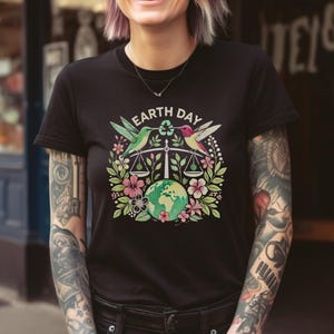 May include: A black t-shirt with a graphic design featuring two hummingbirds perched on a scale with a globe in the center. The text "EARTH DAY" is above the scale. The graphic is surrounded by flowers and leaves.