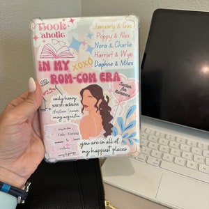 May include: A white and pink book cover with the text "In My Rom-Com Era" and various other text and illustrations. The cover features a woman with brown hair and a pink dress.