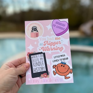 May include: A set of five stickers with a pink background. The stickers feature a book with the text "SMUT SMUT SMUT", a heart with the text "ENEMIES TO LOVERS", a black rectangle with the text "WHEN I DIE DELETE MY KINDLE HISTORY", a cartoon character with the text "LITTLE MISS LOVES TO READ", and a pink banner with the text "You had me at Trigger Warning in my kindle era".