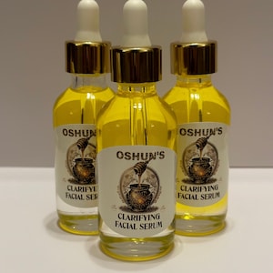 Könnte beinhalten: Drei bernsteinfarbene Glasflaschen mit goldenen Deckeln und Tropfern, gefüllt mit einer gelben Flüssigkeit. Jede Flasche hat ein Etikett mit dem Text "OSHUN'S" und "CLARIFYING FACIAL SERUM".