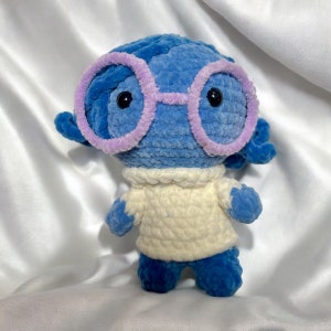 May include: A blue crocheted plush toy with a white crocheted sweater and purple glasses. The toy has a sad expression.