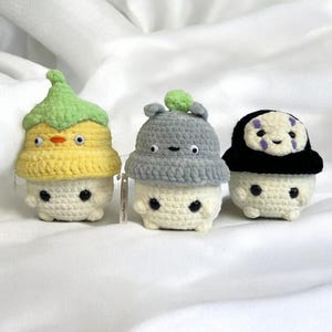 May include: Three crocheted plush toys, each with a different character design. The first is yellow with a green hat, the second is gray with a green hat, and the third is black with a purple face.