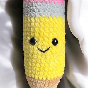 May include: A handmade, smiling crochet pencil in yellow, gray, pink, and tan. The pencil has black button eyes and a stitched smile. The pencil is a soft, plush toy.