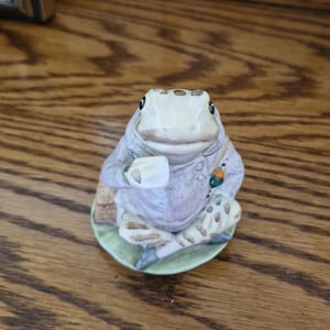 Könnte beinhalten: Eine Keramik-Froschfigur, die auf einem grünen Sockel sitzt. Der Frosch trägt ein lila Hemd und hält eine weiße Tasse in der Hand. Der Frosch hat braune und weiße Flecken auf seinem Körper.