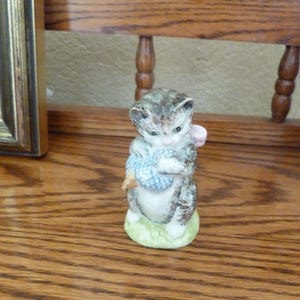 Può includere: Una statuetta in ceramica di un gatto grigio e bianco che tiene una palla blu e bianca. Il gatto ha le orecchie rosa e un fiocco rosa. La statuetta è in piedi su una base verde.