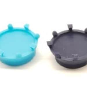 Puede incluir: Dos tapones de botella redondos de plástico. Uno es azul claro y el otro es azul oscuro.