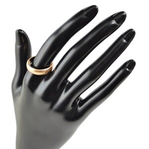 Könnte beinhalten: Ein goldener Bandring mit einem strukturierten Design. Der Ring wird an einer schwarzen Mannequin-Hand getragen.