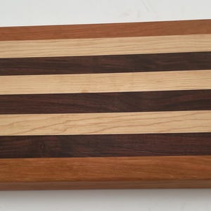 Può includere: Un tagliere rettangolare in legno caratterizzato da strisce orizzontali alternate di legno color marrone chiaro, marrone scuro e marrone rossastro medio. La superficie è liscia e piatta, mettendo in risalto le venature naturali del legno.