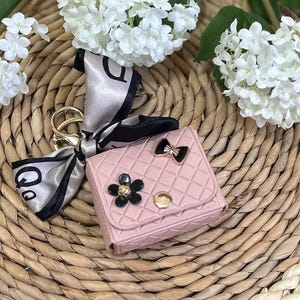 May include: A pink quilted keychain with a black and gold bow, a black flower with a gold center, and a gold button. The keychain is attached to a black and silver scarf.