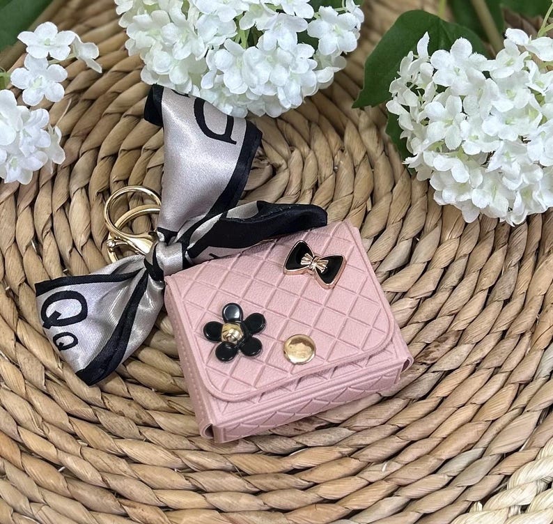May include: A pink quilted keychain with a black and gold bow, a black flower with a gold center, and a gold button. The keychain is attached to a black and silver scarf.