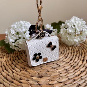 May include: White faux leather handbag keychain with a gold chain, black bow, white flower, and a gold charm with the letter "W".