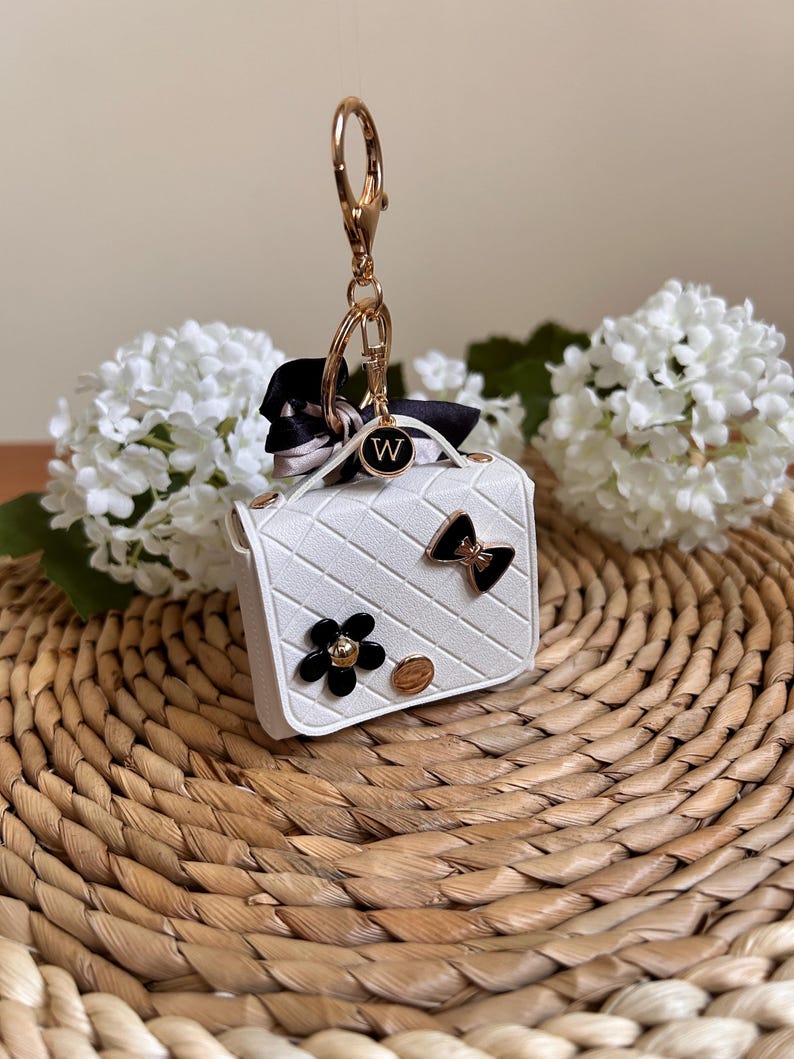 May include: White faux leather handbag keychain with a gold chain, black bow, white flower, and a gold charm with the letter "W".