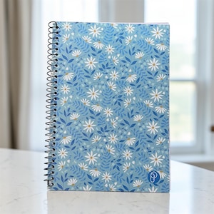 Op de afbeelding: Een spiraalgebonden notitieboek met een lichtblauwe kaft met een bloemenpatroon van witte madeliefjes en blauwe bladeren. Het notitieboek heeft een zwarte spiraalbinding en een klein logo in de rechter benedenhoek. Het notitieboek ligt op een wit oppervlak.