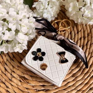 May include: White quilted keychain with a black bow and flower embellishment. The keychain has a gold ring and a black and white patterned fabric attached.