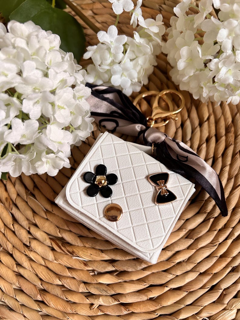 May include: White quilted keychain with a black bow and flower embellishment. The keychain has a gold ring and a black and white patterned fabric attached.