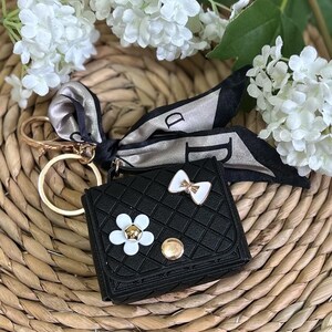 May include: Black faux leather keychain with a small purse design, featuring a white flower and bow embellishment. A gold chain and a black and white patterned scarf are attached.