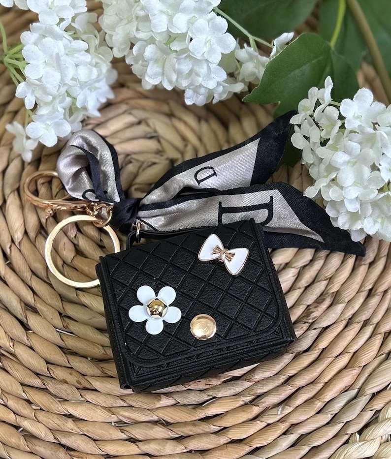 May include: Black faux leather keychain with a small purse design, featuring a white flower and bow embellishment. A gold chain and a black and white patterned scarf are attached.