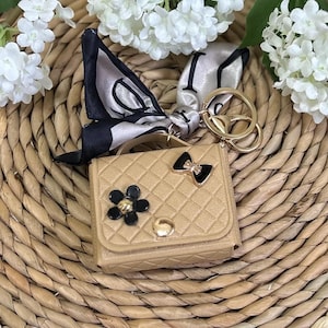 May include: A small, tan faux leather handbag keychain with a black and gold flower, a black bow, and a gold chain with a key ring. The keychain is on a woven straw surface.